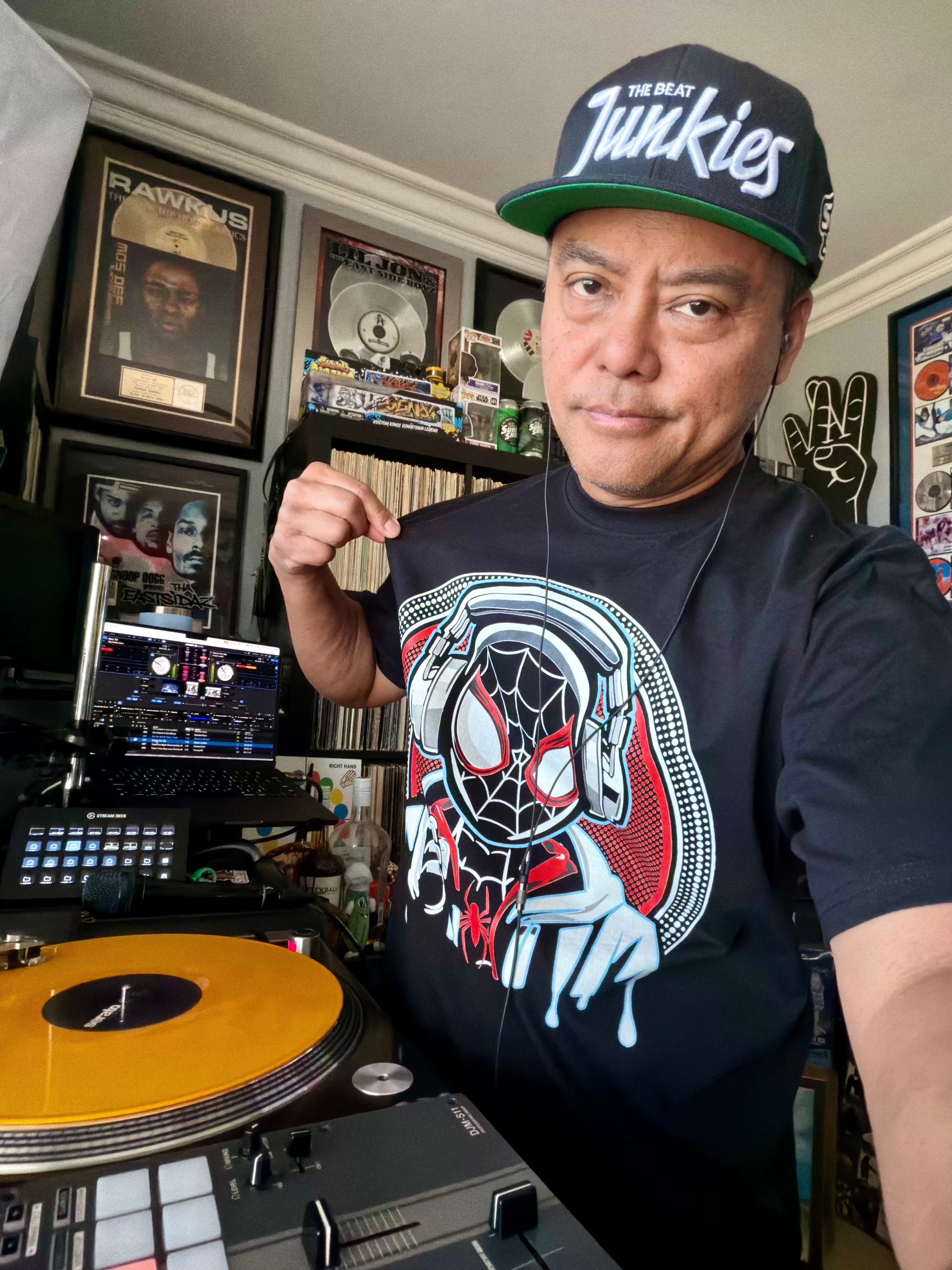 Person wearing a black t-shirt with a graphic design, standing in front of DJ equipment and records.