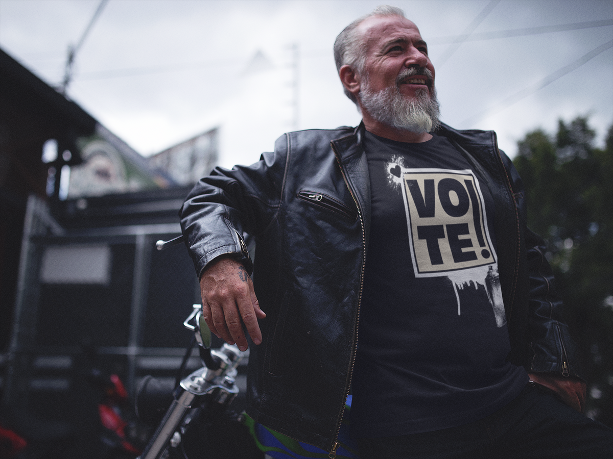 A smiling older man with a gray beard wearing a black leather jacket and a black graphic t-shirt that reads 'VOTE!' in bold white letters, posed outdoors next to a motorcycle.