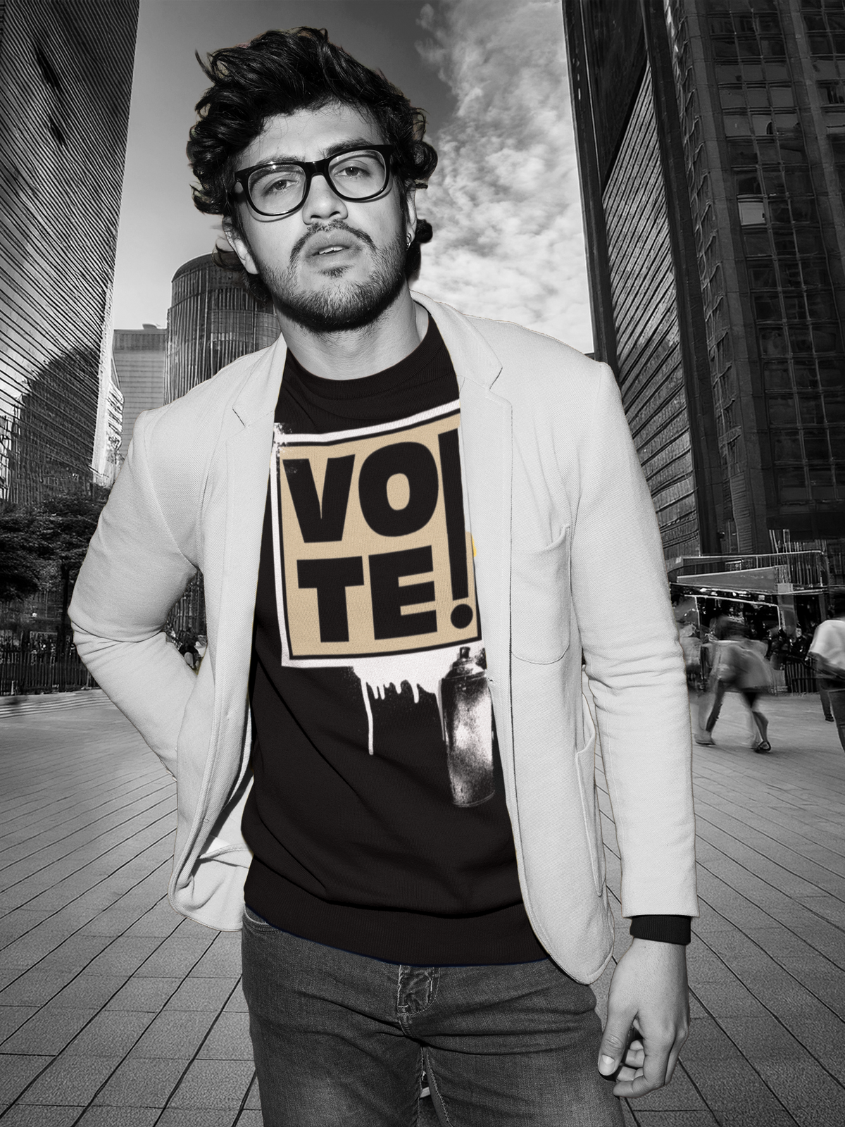 A man wearing a black graphic t-shirt with a beige 'VOTE!' print, styled with a light-colored blazer and dark jeans, set against a blurred black-and-white city street background.