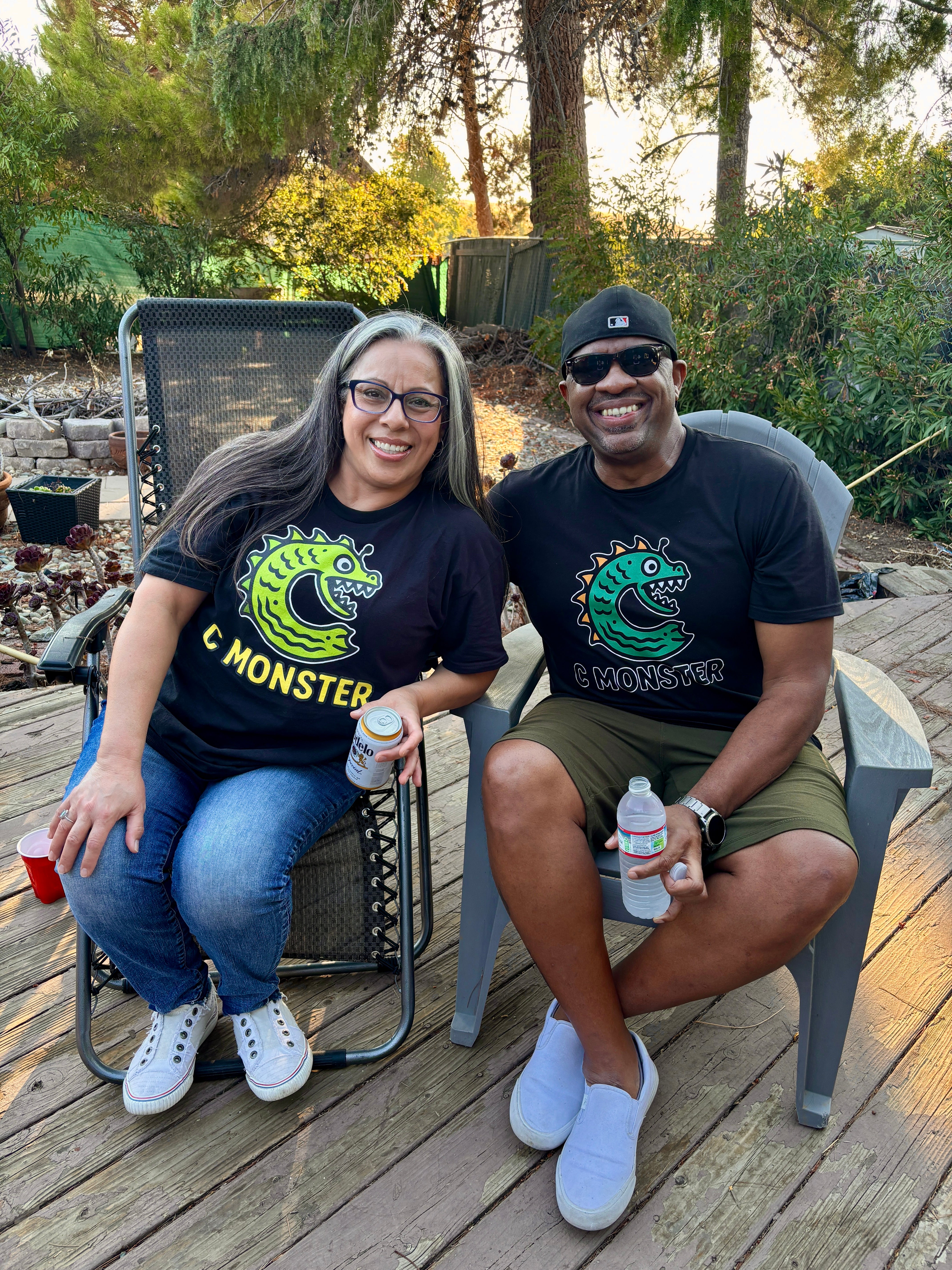 Two people sitting outdoors on a wooden deck, wearing matching black t-shirts with a green logo.